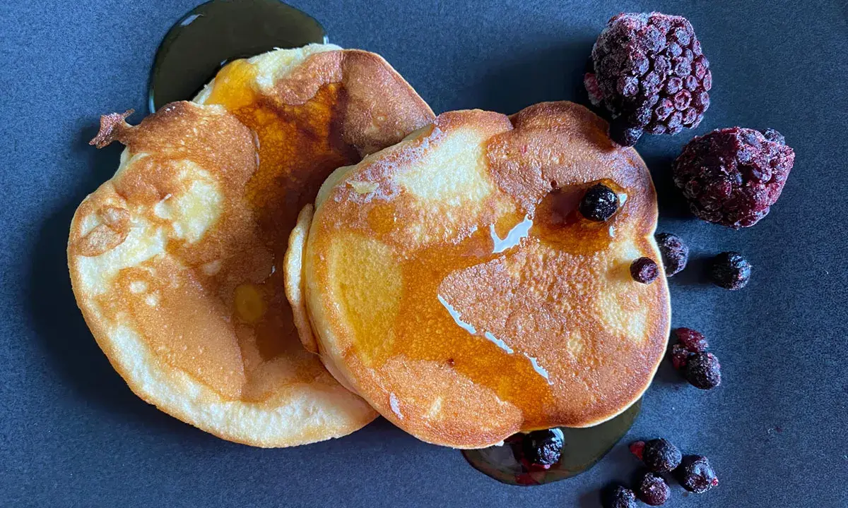 Zwei goldbraune Pancakes ohne Backpulver, beträufelt mit Sirup und garniert mit gefrorenen Beeren auf dunklem Teller.