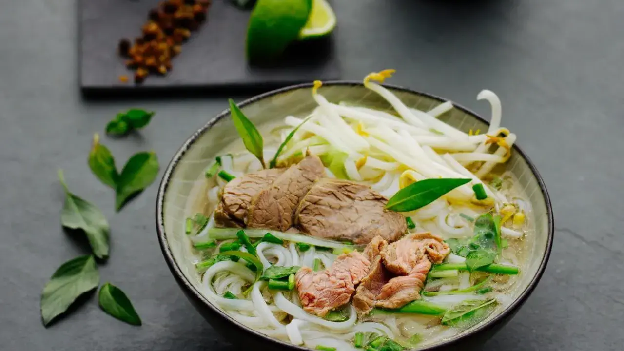 Eine dampfende Schüssel Pho Suppe mit zarten Rindfleischscheiben, Reisnudeln, Sojasprossen und frischen Kräutern. Ein Hauch von Limette und Gewürzen im Hintergrund.