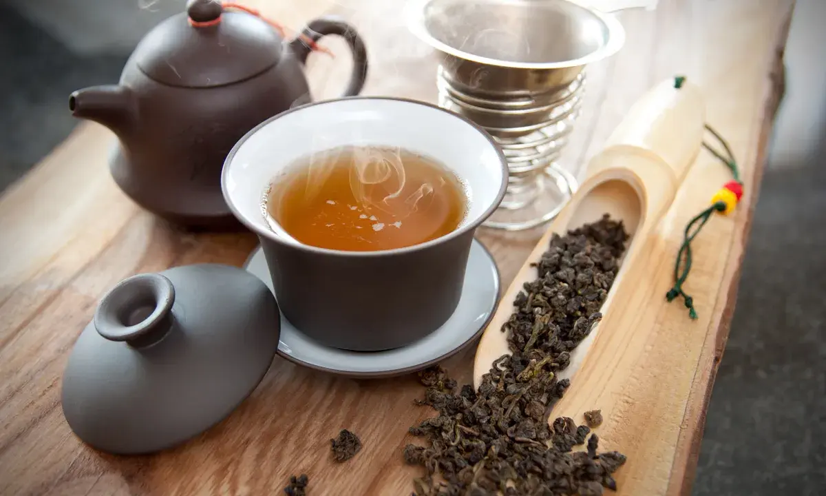 Dampf steigt aus einer Tasse Oolong Tee auf. Daneben liegen lose Teeblätter in einer Holzschaufel. So schmeckt Oolong Tee: mild, blumig, leicht süßlich.