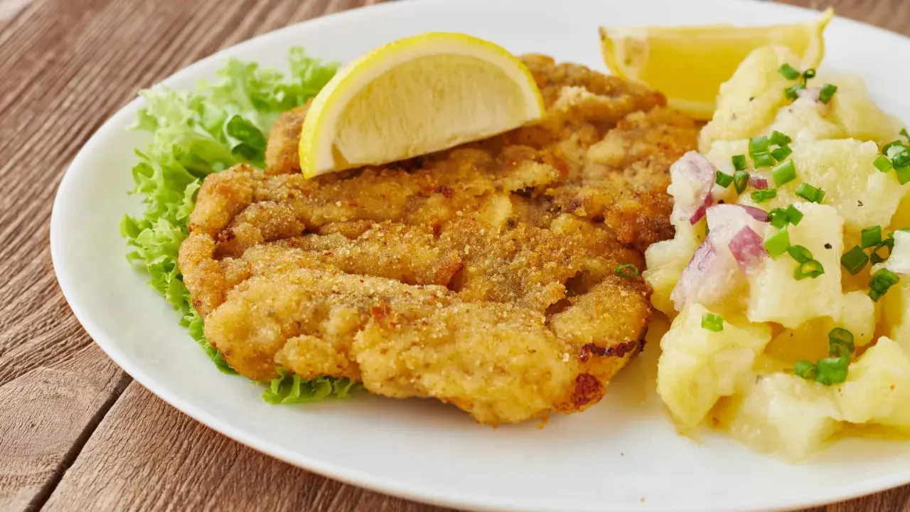 Ein knuspriges Wiener Schnitzel mit Zitrone, Salat und Kartoffelsalat auf einem weißen Teller.