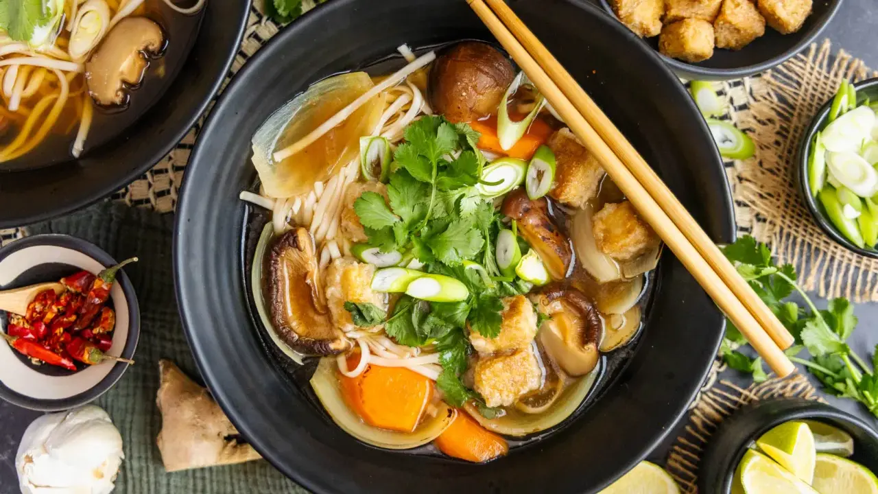 Herzhaftes Pho Chay mit Udon-Nudeln, Shiitake-Pilzen, Tofu und frischem Koriander. Daneben kleine Schälchen mit Chili, Lauchzwiebeln und Limettenspalten.