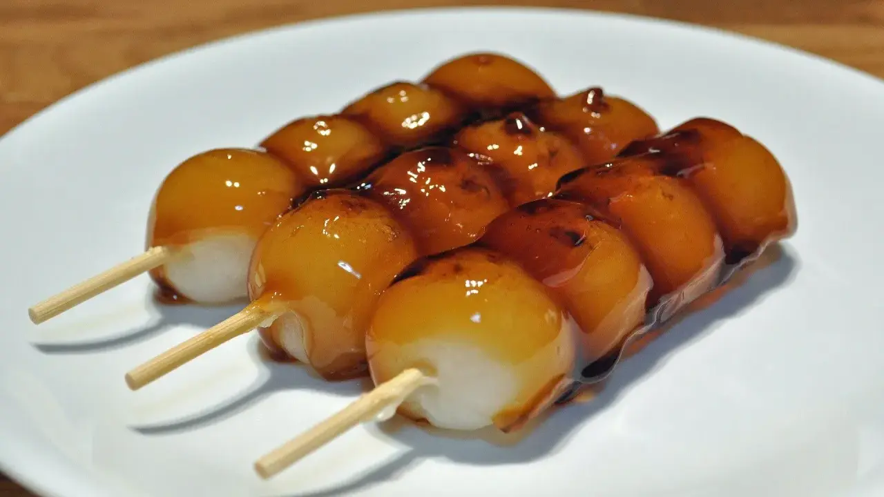 Drei Spieße mit süßen, glasierten japanischen Desserts, die auf einem weißen Teller liegen.