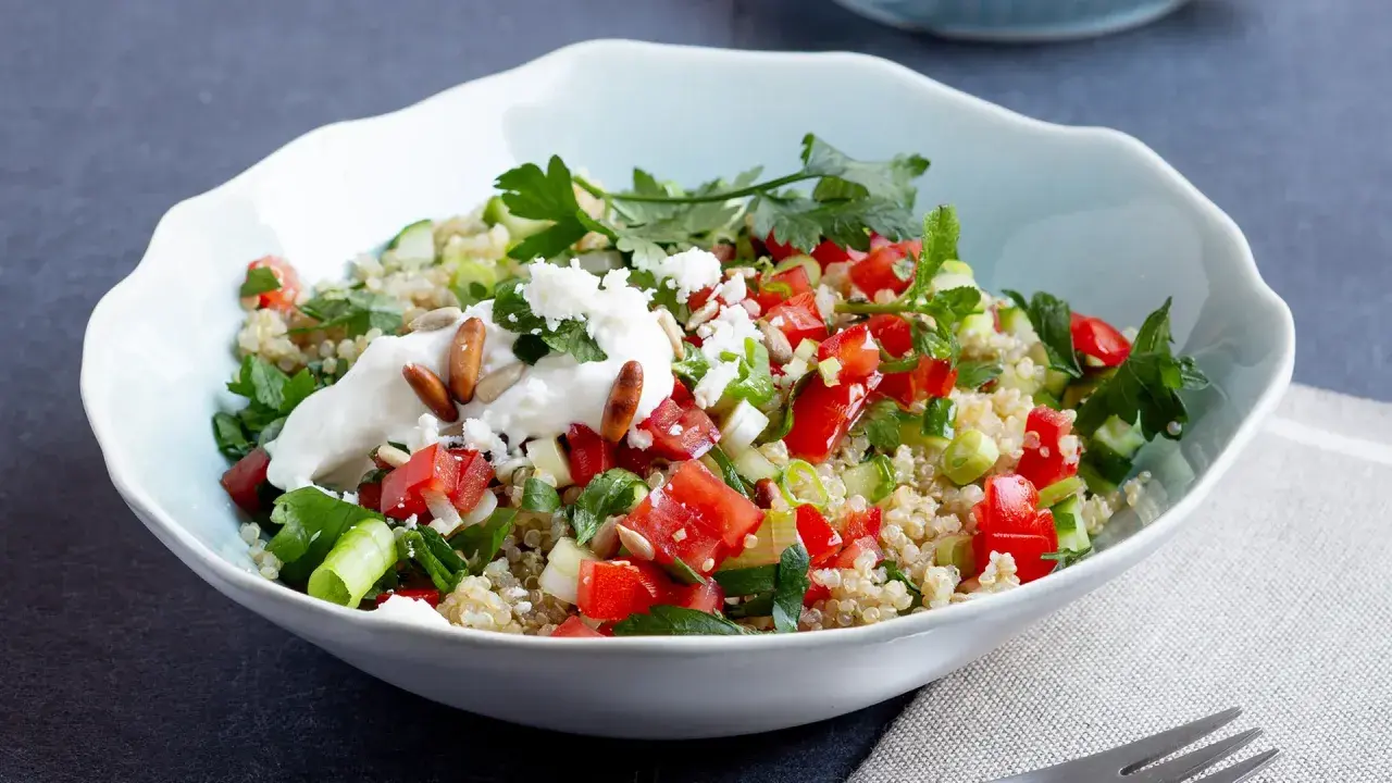 Frischer Quinoa-Salat mit Feta, Tomaten, Gurken und Kräutern, garniert mit Joghurt und Pinienkernen.