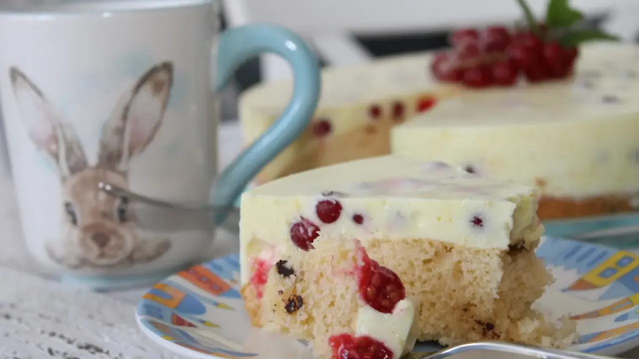 Ein Stück Johannisbeertorte mit Joghurt, dekoriert mit frischen roten Johannisbeeren. Daneben eine Tasse mit Hasenmotiv.