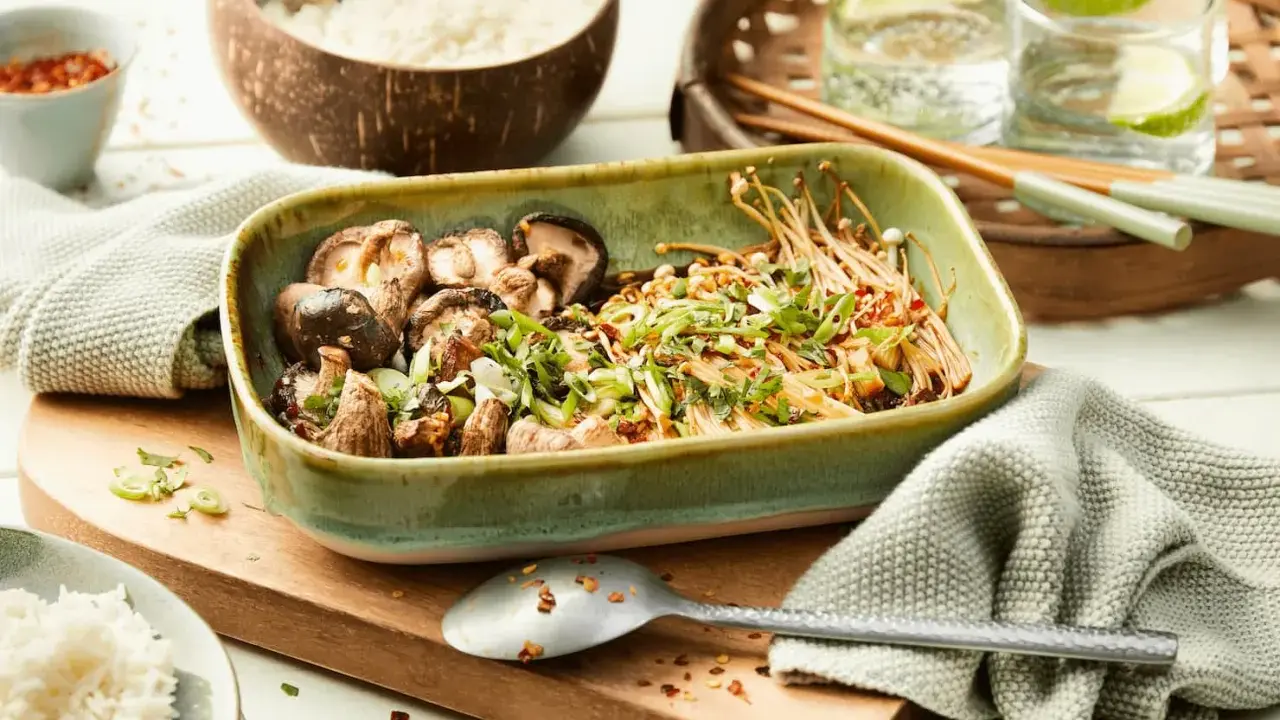 Schüssel mit gebratenen asiatischen Pilzen, garniert mit Frühlingszwiebeln und Koriander, daneben Reis und Chilis.