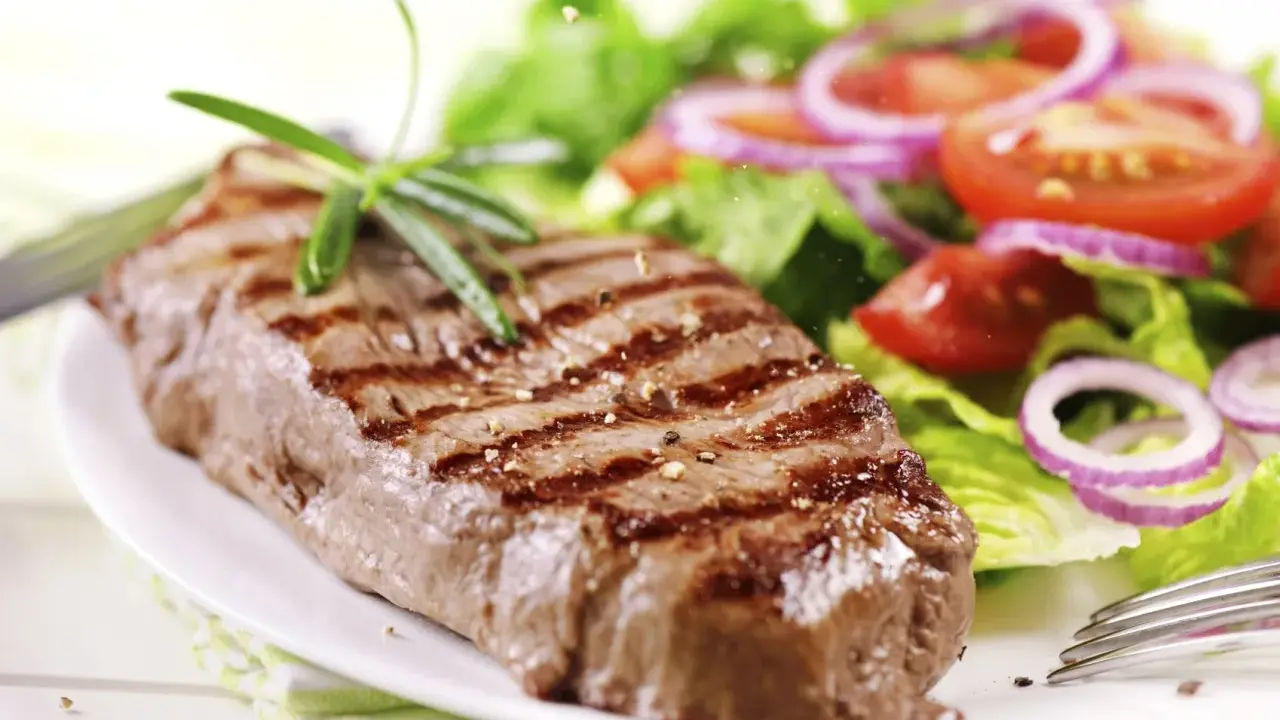 Ein saftiges Steak mit Grillstreifen, garniert mit Rosmarin, neben einem frischen Salat aus knackigem Grün, Tomaten und roten Zwiebelringen. Ein perfekter Steak Salat.