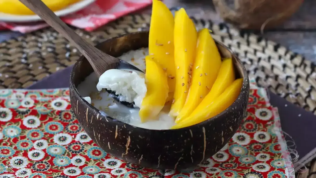Ein Schälchen mit Mango Sticky Rice, garniert mit frischen Mangoscheiben und Sesam. Ein Löffel hebt cremigen Klebreis mit Kokosmilch hervor.