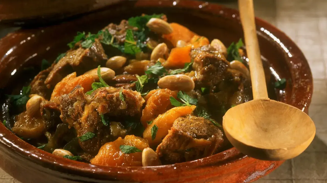 Marokkanisches Lamm mit Aprikosen, Mandeln und frischer Minze in einem traditionellen Tajine-Topf, serviert mit einem Holzlöffel.