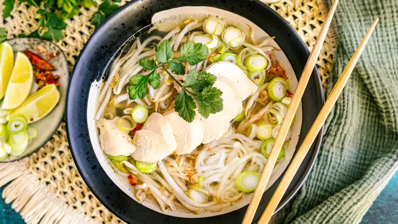 Eine dampfende Schüssel Pho Ga mit zarten Hühnerbrustscheiben, Reisnudeln, Sojasprossen und Frühlingszwiebeln. Serviert mit Limettenspalten und Chili.