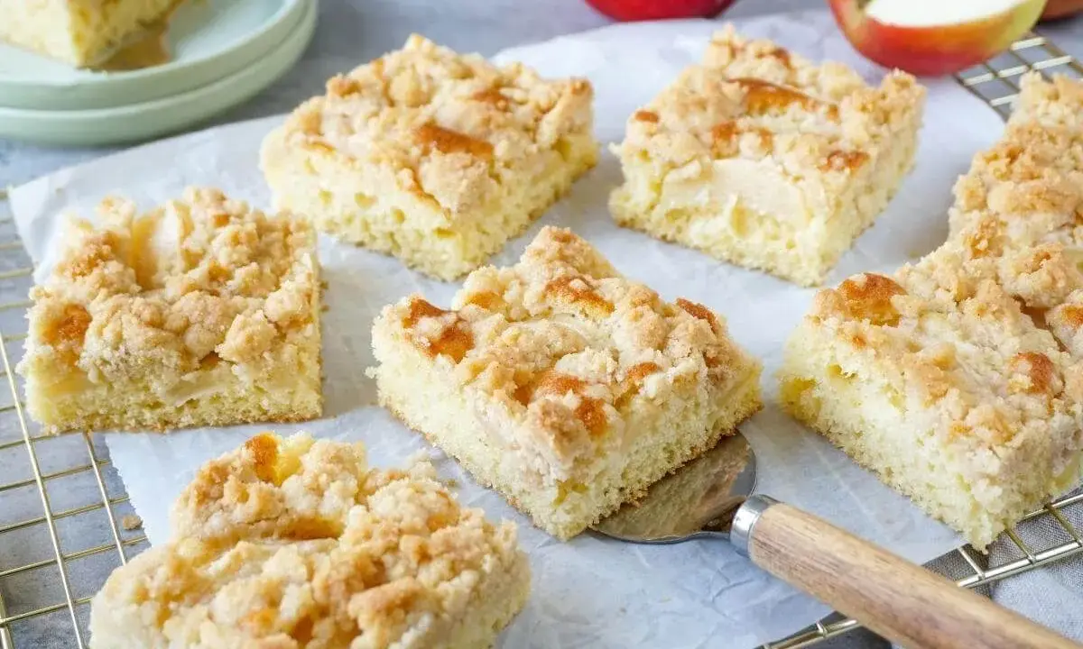 Saftige Stücke vom besten Apfelkuchen mit Streusel, frisch gebacken und bereit zum Genießen. Äpfel liegen daneben.