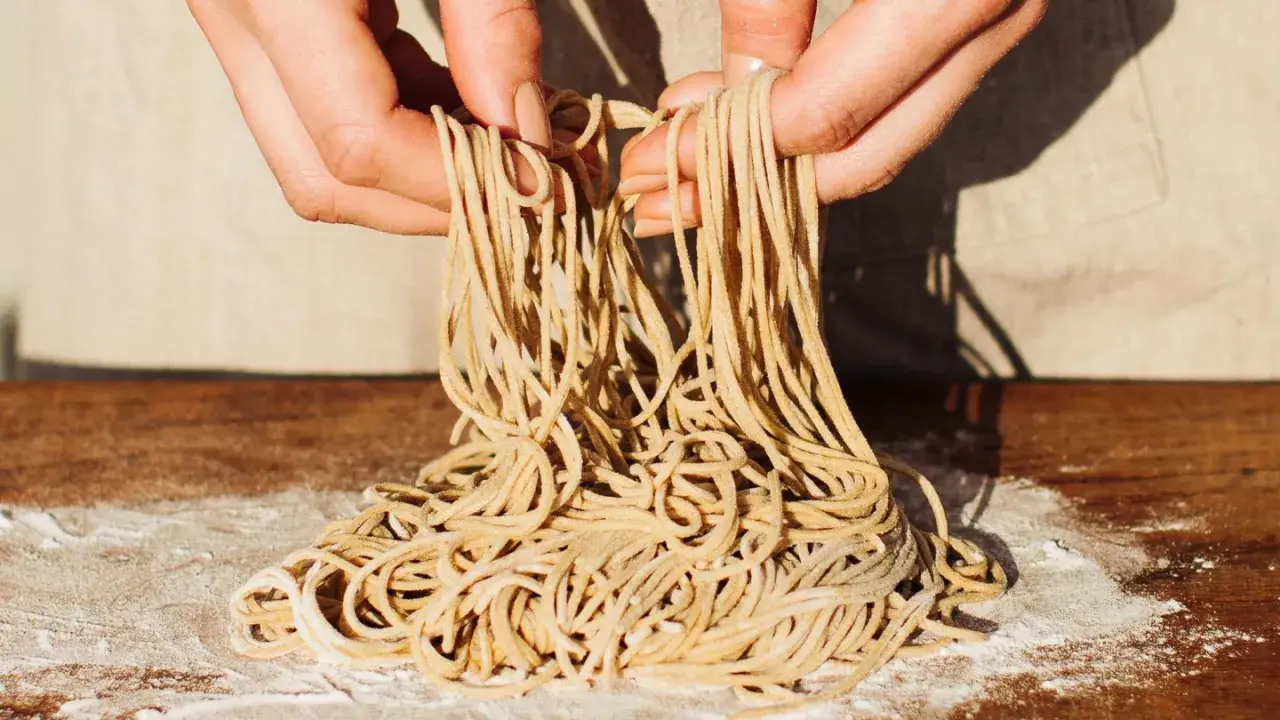 Hände halten frisch gemachte Ramen-Nudeln über einem Holztisch mit Mehl. So einfach kann ramen selber machen sein!