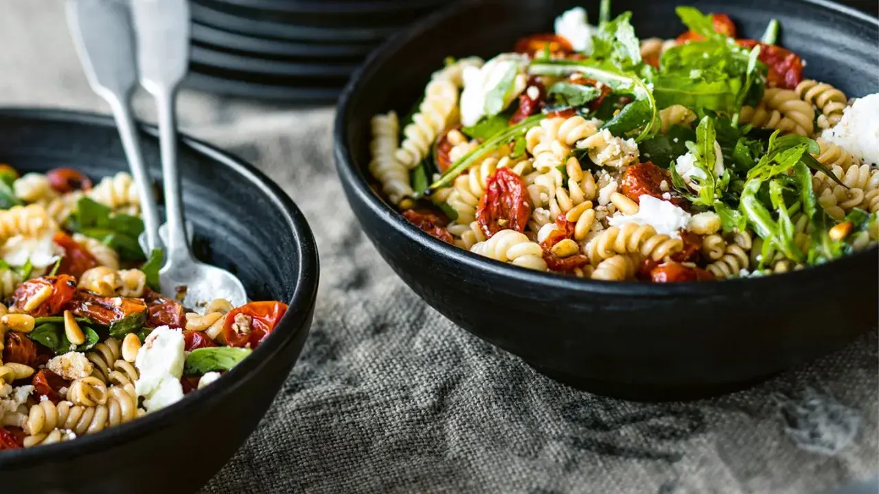 Zwei Schalen mit einem gesunden Nudelsalat aus Spiralnudeln, getrockneten Tomaten, Pinienkernen, Rucola und Feta.