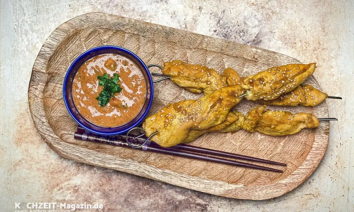 Saftige Satay Chicken Spieße mit Sesam, serviert mit Erdnusssauce und Stäbchen auf einem Holztablett.