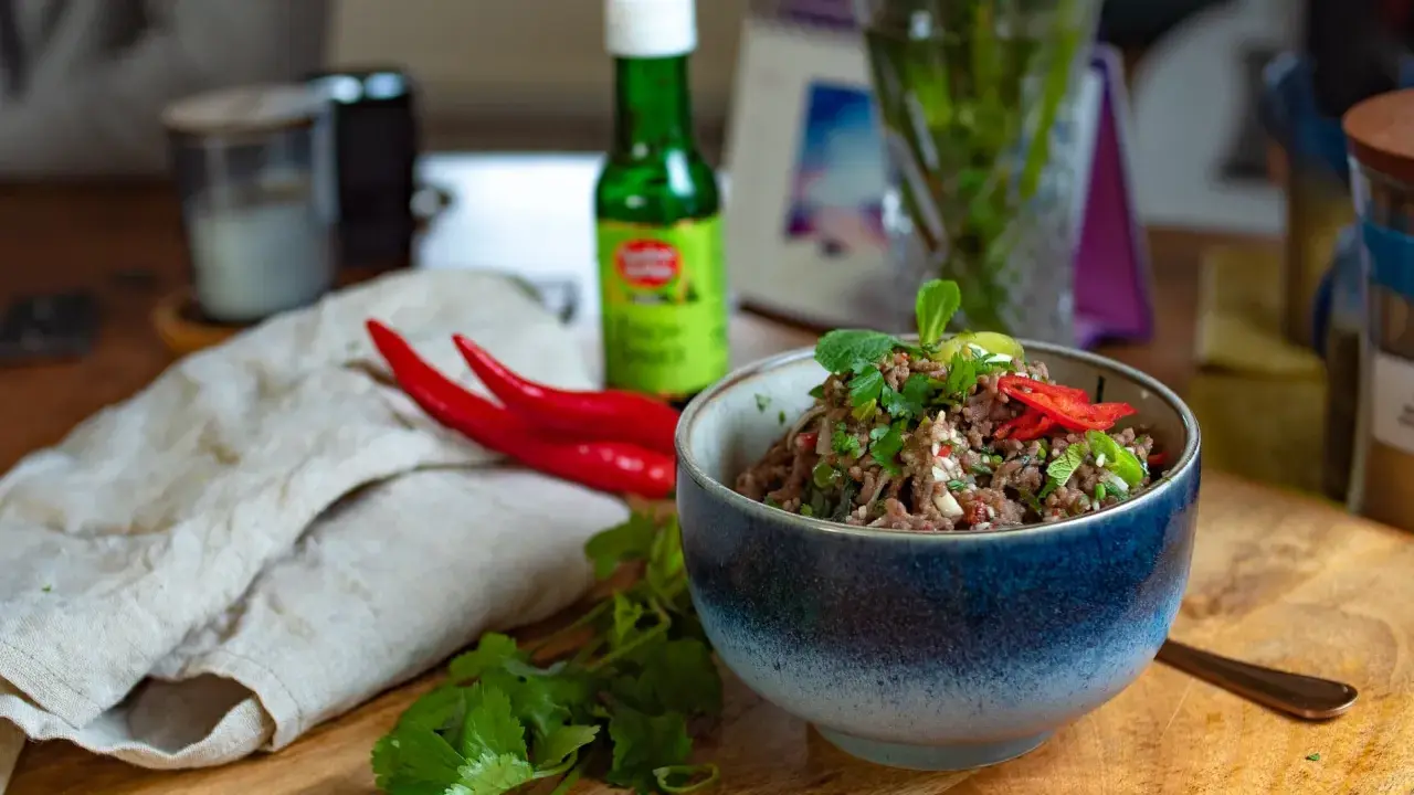 Würziges Laab Moo, ein thailändischer Hackfleischsalat, garniert mit frischer Minze und Chili. Daneben liegen rote Chilis und Koriander.