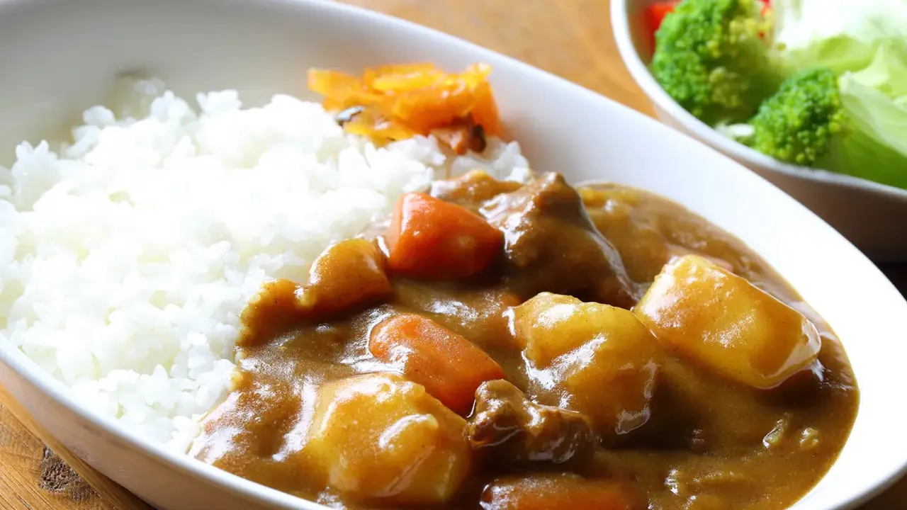 Ein Teller mit dampfendem Reis und herzhaftem Curry, das japanische Nationalgericht, mit zarten Fleischstücken, Kartoffeln und Karotten. Daneben ein kleiner Salat.