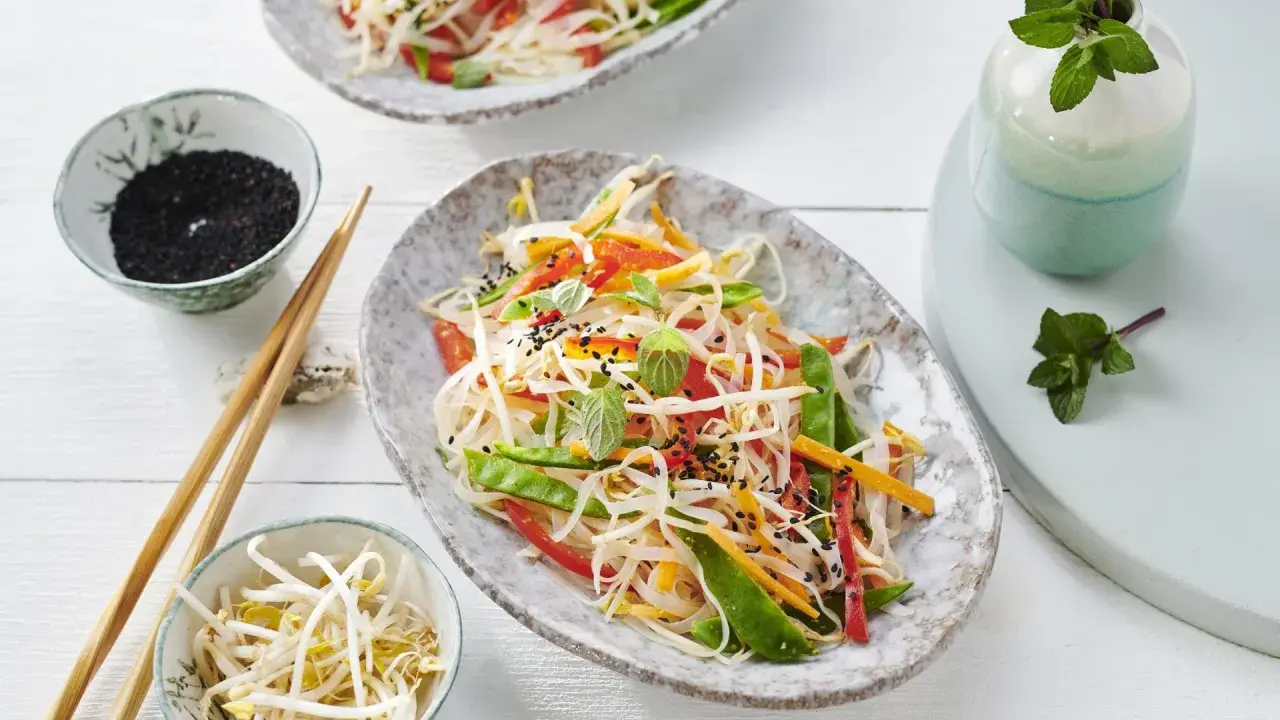 Frischer Glasnudelsalat mit knackigem Gemüse, garniert mit Sesam und Minze. Daneben Schüsselchen mit Sprossen und schwarzem Sesam.