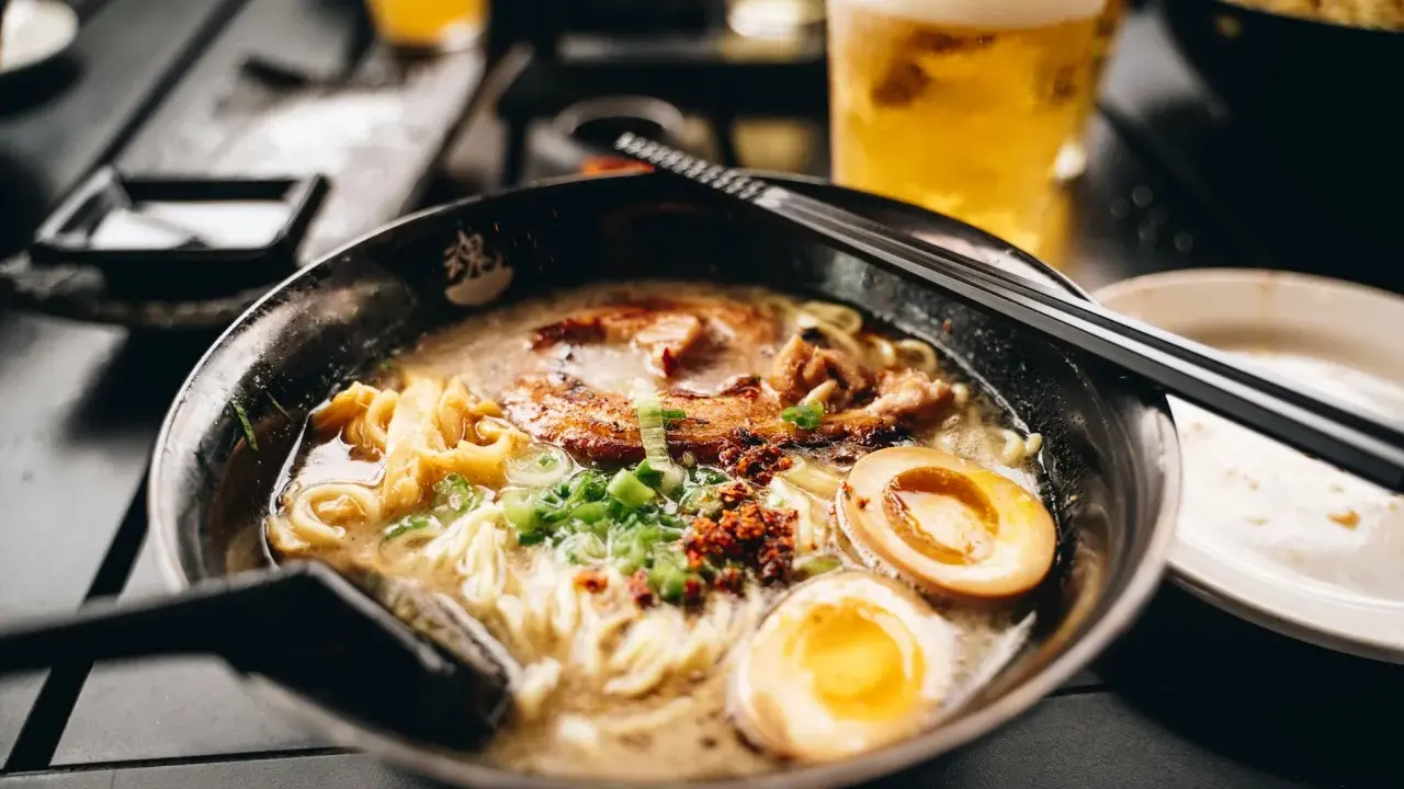 Eine dampfende Schüssel mit den besten Ramen-Bowls, gefüllt mit Nudeln, Schweinefleisch, Ei und Frühlingszwiebeln. Daneben ein Glas Bier.