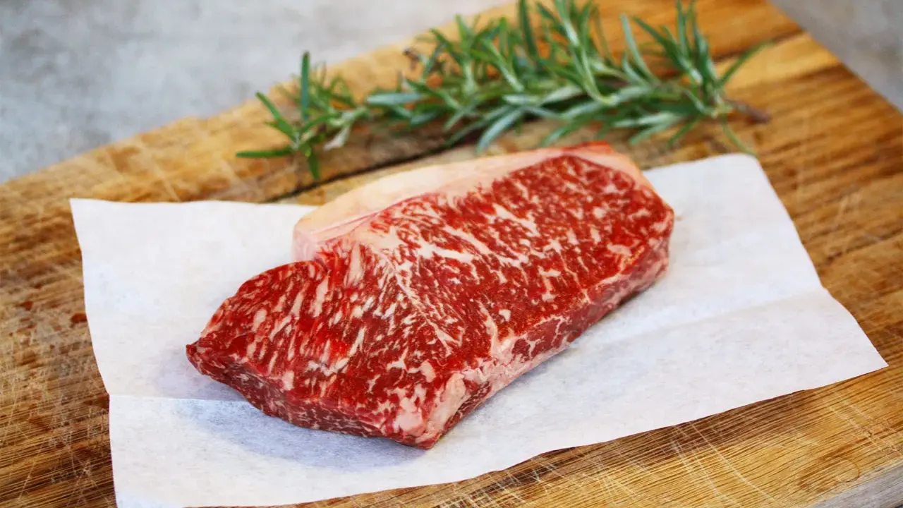 Ein rohes Stück Wagyu-Fleisch mit feiner Marmorierung liegt auf Backpapier auf einem Holzbrett. Ein Rosmarinzweig liegt daneben.