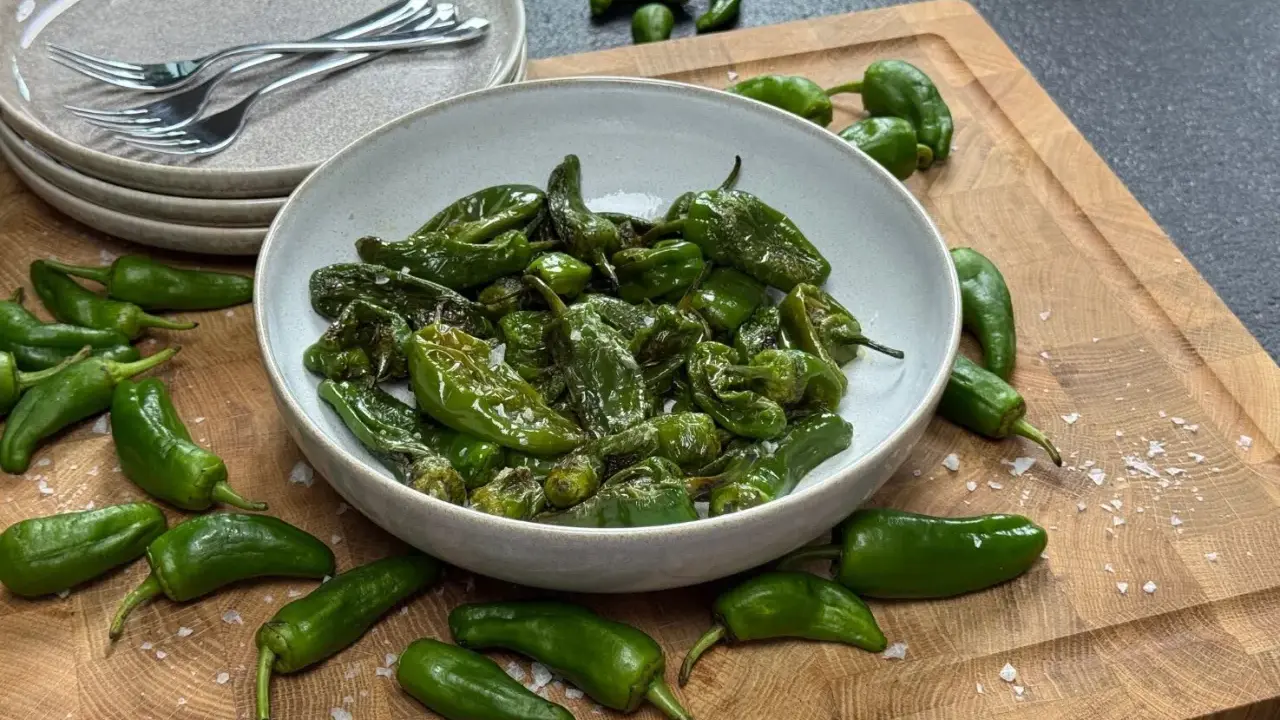 Schüssel mit gebratenen Pimientos de Padrón, bestreut mit grobem Salz. Einige rohe Paprika liegen daneben auf einem Holzbrett.