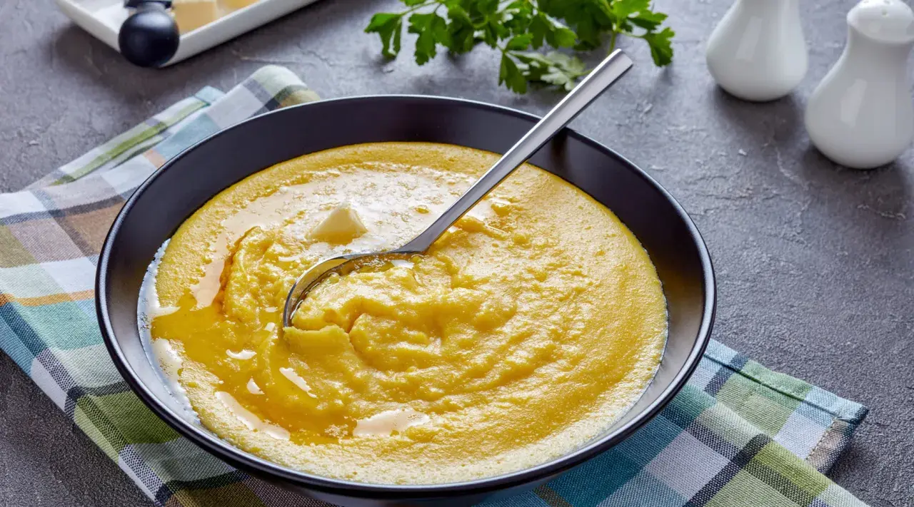 Eine Schüssel cremiger Polenta mit einem Stück Butter und einem Löffel. Was ist Polenta? Ein herzhaftes Maisgericht, das warm serviert wird.