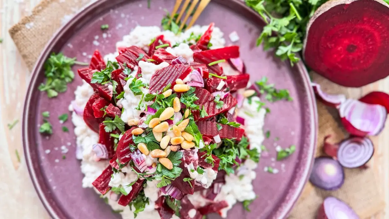 Roter Bete Salat mit cremigem Käse, Pinienkernen und frischer Petersilie auf einem lila Teller.