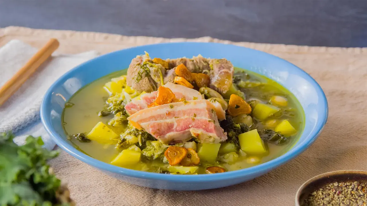Herzhafter spanischer Eintopf mit Fleisch, Kartoffeln, Grünkohl und Karotten in einer blauen Schüssel.