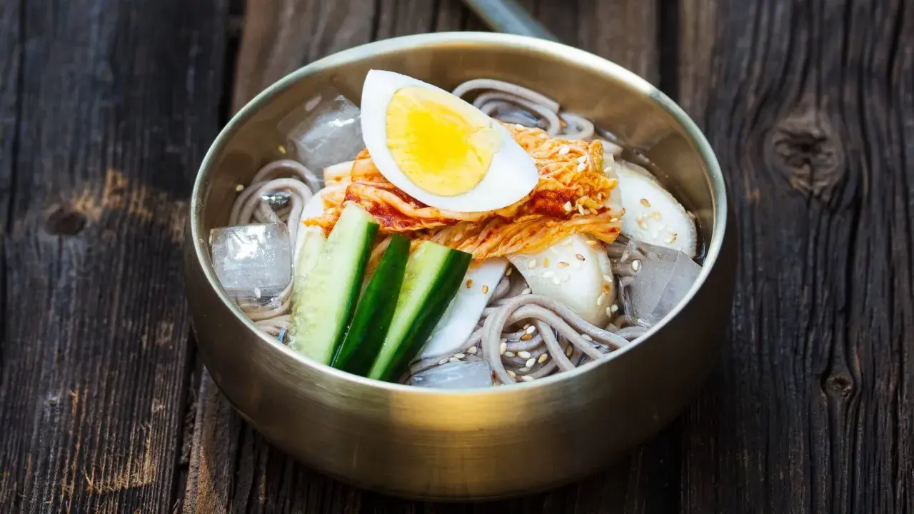 Erfrischendes Mul Naengmyeon mit Buchweizennudeln, Kimchi, Gurke, Ei und Eiswürfeln in einer bronzenen Schüssel auf Holzuntergrund.