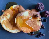 Zwei goldbraune Pancakes ohne Backpulver, beträufelt mit Sirup und garniert mit gefrorenen Beeren auf dunklem Teller.