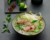 Eine dampfende Schüssel Pho Suppe mit zarten Rindfleischscheiben, Reisnudeln, Sojasprossen und frischen Kräutern. Ein Hauch von Limette und Gewürzen im Hintergrund.