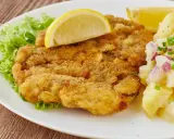 Ein knuspriges Wiener Schnitzel mit Zitrone, Salat und Kartoffelsalat auf einem weißen Teller.