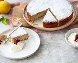 Ein saftiger Ricotta Mandel Kuchen, bestäubt mit Puderzucker, serviert mit einer Kugel Sahne und roter Grütze.