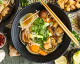 Herzhaftes Pho Chay mit Udon-Nudeln, Shiitake-Pilzen, Tofu und frischem Koriander. Daneben kleine Schälchen mit Chili, Lauchzwiebeln und Limettenspalten.