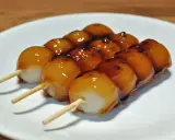 Drei Spieße mit süßen, glasierten japanischen Desserts, die auf einem weißen Teller liegen.