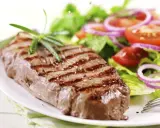 Ein saftiges Steak mit Grillstreifen, garniert mit Rosmarin, neben einem frischen Salat aus knackigem Grün, Tomaten und roten Zwiebelringen. Ein perfekter Steak Salat.