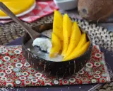 Ein Schälchen mit Mango Sticky Rice, garniert mit frischen Mangoscheiben und Sesam. Ein Löffel hebt cremigen Klebreis mit Kokosmilch hervor.