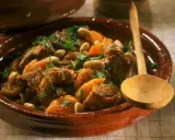 Marokkanisches Lamm mit Aprikosen, Mandeln und frischer Minze in einem traditionellen Tajine-Topf, serviert mit einem Holzlöffel.