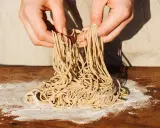 Hände halten frisch gemachte Ramen-Nudeln über einem Holztisch mit Mehl. So einfach kann ramen selber machen sein!