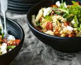 Zwei Schalen mit einem gesunden Nudelsalat aus Spiralnudeln, getrockneten Tomaten, Pinienkernen, Rucola und Feta.