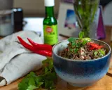 Würziges Laab Moo, ein thailändischer Hackfleischsalat, garniert mit frischer Minze und Chili. Daneben liegen rote Chilis und Koriander.