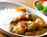 Ein Teller mit dampfendem Reis und herzhaftem Curry, das japanische Nationalgericht, mit zarten Fleischstücken, Kartoffeln und Karotten. Daneben ein kleiner Salat.