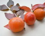 Drei leuchtend orangefarbene Kaki-Früchte liegen neben herbstlichen Blättern.