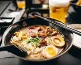 Eine dampfende Schüssel mit den besten Ramen-Bowls, gefüllt mit Nudeln, Schweinefleisch, Ei und Frühlingszwiebeln. Daneben ein Glas Bier.