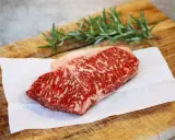 Ein rohes Stück Wagyu-Fleisch mit feiner Marmorierung liegt auf Backpapier auf einem Holzbrett. Ein Rosmarinzweig liegt daneben.