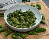 Schüssel mit gebratenen Pimientos de Padrón, bestreut mit grobem Salz. Einige rohe Paprika liegen daneben auf einem Holzbrett.