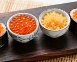 Zwei Sushi-Rollen mit Lachsrogen und eine Schale mit Masago, dem Rogen von Capelin-Fischen, daneben eine Schale mit Lachsrogen.