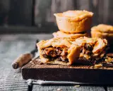 Herzhafte Pasteten mit Fleischfüllung auf einem Holzbrett. Eine Pastete ist aufgeschnitten und zeigt den Inhalt.