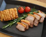 Knuspriger asiatischer Schweinebauch, in Scheiben geschnitten, mit Rosmarin und Kirschtomaten auf einer Gusseisenplatte serviert.