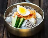Erfrischendes Mul Naengmyeon mit Buchweizennudeln, Kimchi, Gurke, Ei und Eiswürfeln in einer bronzenen Schüssel auf Holzuntergrund.