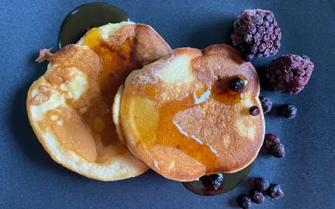 Zwei goldbraune Pancakes ohne Backpulver, beträufelt mit Sirup und garniert mit gefrorenen Beeren auf dunklem Teller.