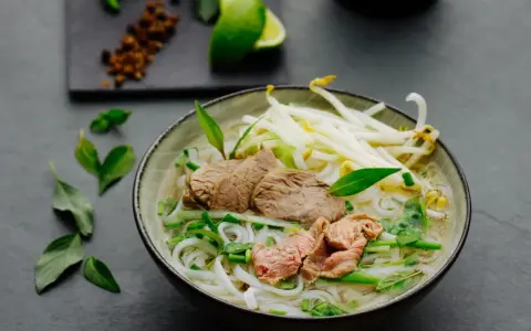 Eine dampfende Schüssel Pho Suppe mit zarten Rindfleischscheiben, Reisnudeln, Sojasprossen und frischen Kräutern. Ein Hauch von Limette und Gewürzen im Hintergrund.